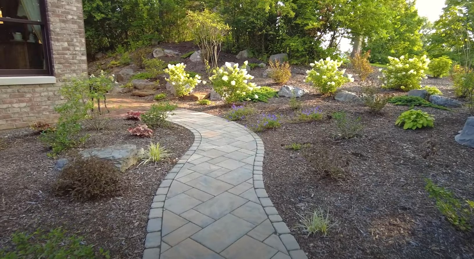 Hardscape walkway installation integrated with natural landscape design Curved stone pathway through professionally designed landscape with flowering shrubs and natural boulder accents